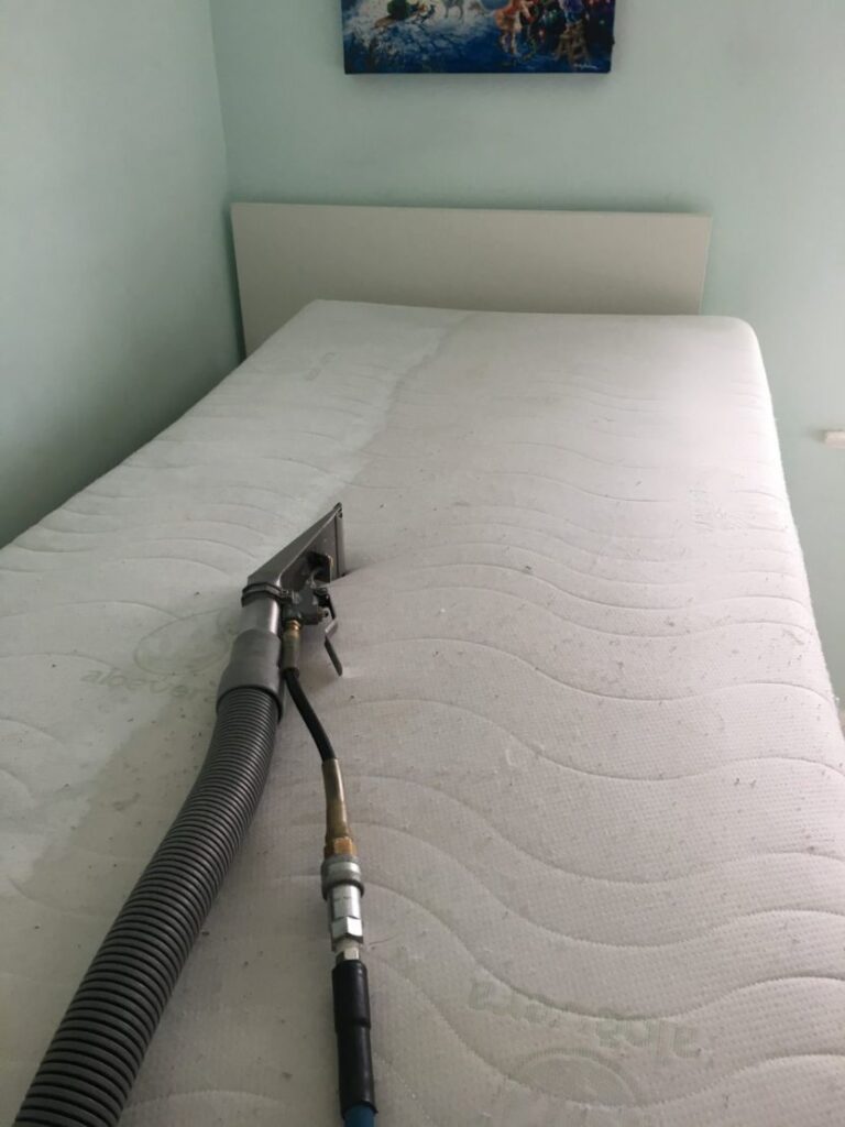 Bed with a white mattress and a cleaning wand attached to a hose resting on top in a light-blue room.