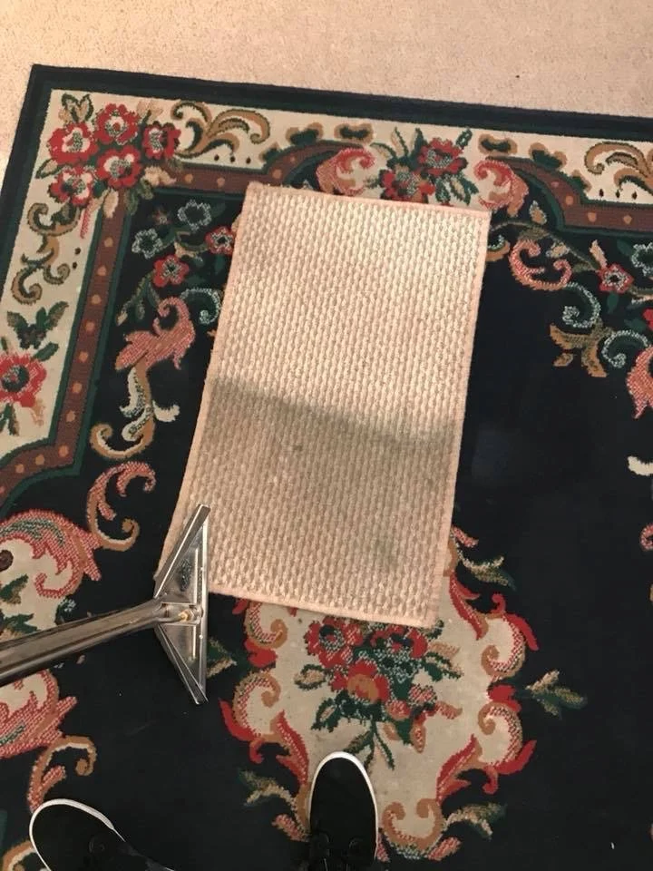 Top-down view of a beige woven rectangular rug lying on a dark ornate oriental rug; a metal tool sits at the lower left and a black sneaker is visible at the bottom edge.