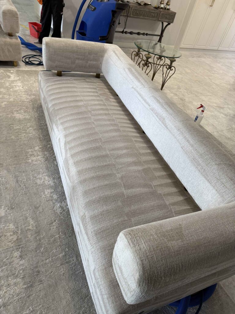 Beige sectional sofa being cleaned in a living room; a blue carpet cleaning machine and hoses are visible in the background